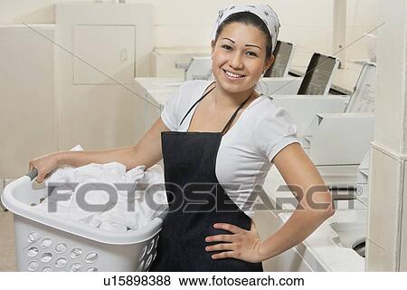 Stock Photo - Portrait of a happy young female employee carrying laundry basket in Laundromat. Fotosearch