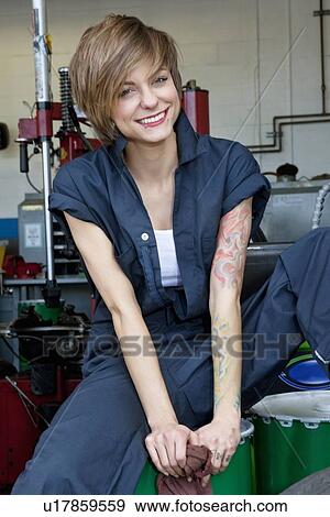Stock Photo - Portrait of a happy young female mechanic sitting on oil drum in car workshop. Fotosearch