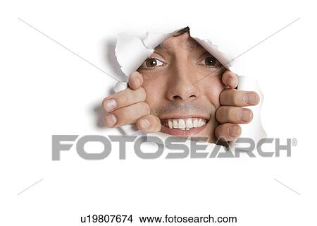 Picture - Portrait of a happy young Hispanic man peeking from ripped paper hole. Fotosearch