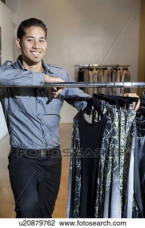Stock Image - Portrait of a happy young male standing by clothes rack in fashion store. Fotosearch