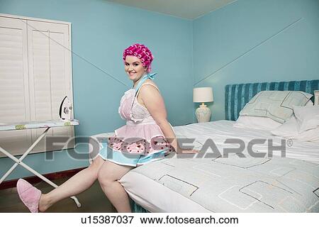 Stock Photo - Portrait of a happy young woman sitting on bed with ironing board in background. Fotosearch