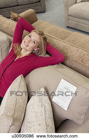 Stock Photograph - Portrait of a happy young woman with hands behind head relaxing on sofa in furniture store. Fotosearch