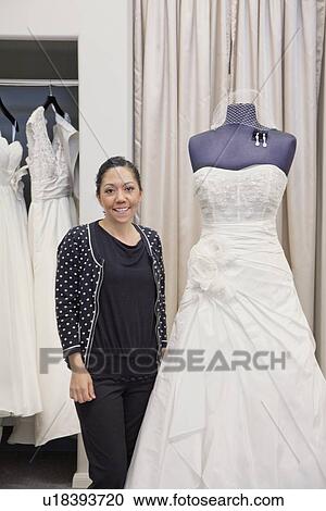 Stock Image - Portrait of a mature employee standing by elegant wedding dress in bridal store. Fotosearch