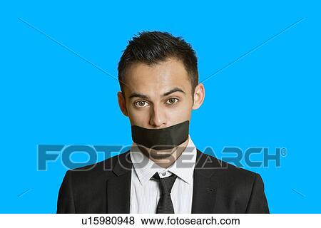 Stock Photo - Portrait of a young businessman with adhesive tape on mouth over colored background. Fotosearch
