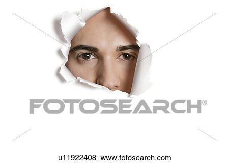 Stock Photo - Portrait of a young Caucasian man peeking from ripped paper hole. Fotosearch