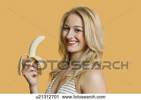 Stock Image - Portrait of a young woman eating banana over colored background. Fotosearch