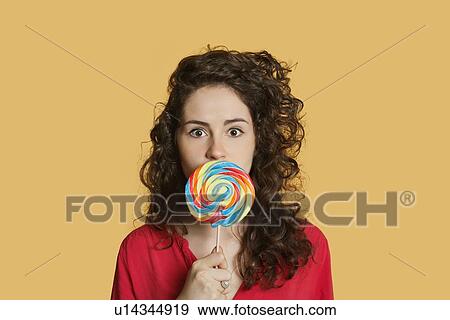 Portrait of a young woman holding lollipop in front of face over colored background View Large Photo Image Stock Photo - Portrait of a young woman holding lollipop in front of face over colored background. Fotosearch