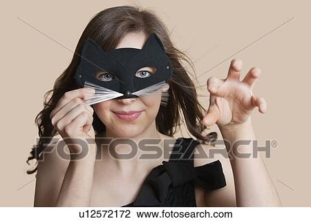 Stock Image - Portrait of a young woman looking through eye mask imitating as cat over colored background. Fotosearch