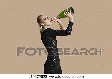 Stock Photo - Portrait of beautiful young woman drinking champagne from bottle over colored background. Fotosearch