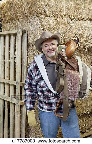 Stock Image - Portrait of happy cowboy carrying riding tack on shoulder. Fotosearch