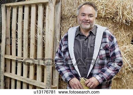 Stock Image - Portrait of happy mature male standing in front of hay stack. Fotosearch