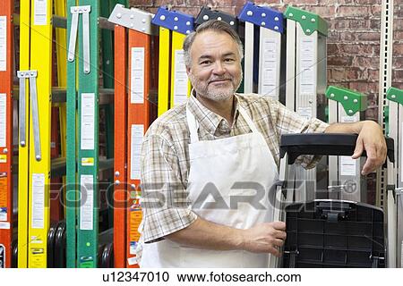 Stock Image - Portrait of happy mature store clerk standing by multicolored ladders in hardware shop. Fotosearch