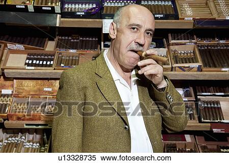 Stock Photography - Portrait of mature tobacco shop owner smoking cigar in store. Fotosearch