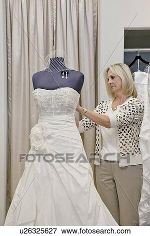 Stock Photo - Senior female owner adjusting wedding dress on mannequin in bridal store. Fotosearch