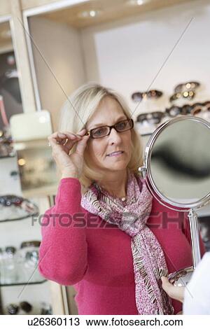 Senior woman trying glasses while looking into hand mirror in store View Large Photo Image Stock Image - Senior woman trying glasses while looking into hand mirror in store. Fotosearch