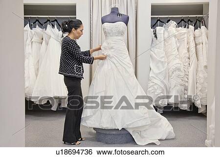 Stock Photograph - Side view of a mature employee adjusting elegant wedding dress in bridal store. Fotosearch