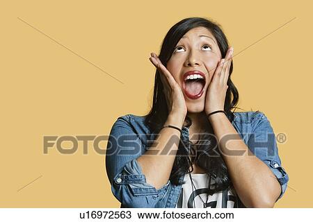 Stock Image - Surprised young woman with head in hands looking up over colored background. Fotosearch