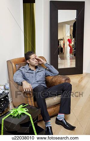 Stock Image - Tired young man sitting on armchair in fashion store. Fotosearch