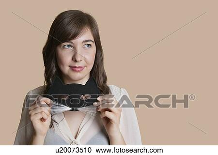 Stock Image - Young woman holding eye mask while looking away over colored background. Fotosearch