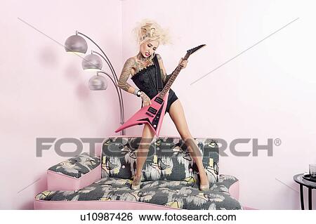 Stock Photograph - Young woman in corset playing guitar while standing on furniture. Fotosearch