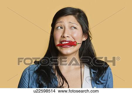Picture - Young woman with red chili pepper in mouth over colored background. Fotosearch