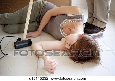Stock Image - Electric shock. Woman lying unconscious on the floor after receiving an electric shock. A man is moving the source of the shock away from the woman. He is using a wooden stick and standing on a book to insulate himself against the electricity.. Fotosearch