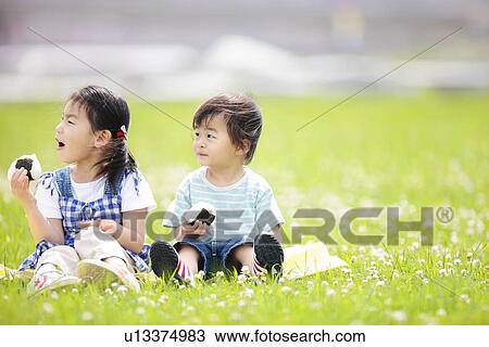 Stock Image - Girl and Boy Sitting on Grass and Eating Rice Ball. Fotosearch