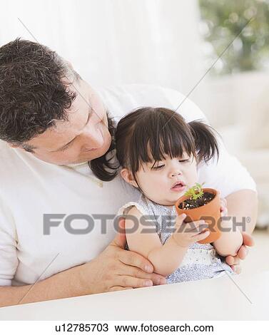 Stock Image - Baby girl (12-17 months) with father watching seedling in pot. Fotosearch