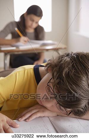 Stock Image - Boy (10-11) napping on desk with girl (12-13) in background. Fotosearch