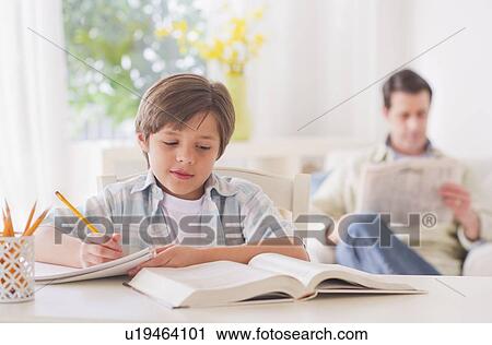 Stock Image - Boy (10-11 years) doing homework at table with father sitting in background. Fotosearch