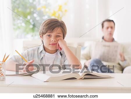 Stock Image - Boy (10-11 years) doing homework at table with father sitting in background. Fotosearch