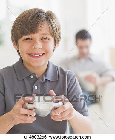 Stock Photograph - Boy (10-11 years) playing video game with father sitting in background. Fotosearch