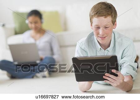 Boy (16-17) lying on floor using tablet pc, girl (14-15) with laptop in background View Large Photo Image Stock Photo - Boy (16-17) lying on floor using tablet pc, girl (14-15) with laptop in background. Fotosearch
