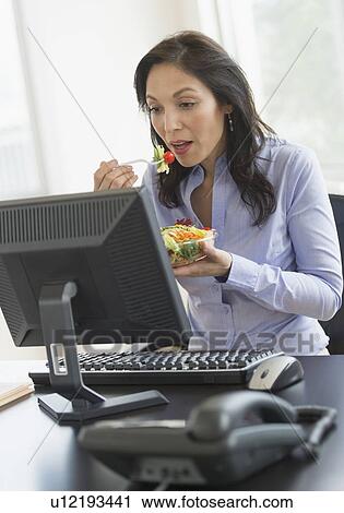 Stock Image - Business woman working on computer and eating salad. Fotosearch