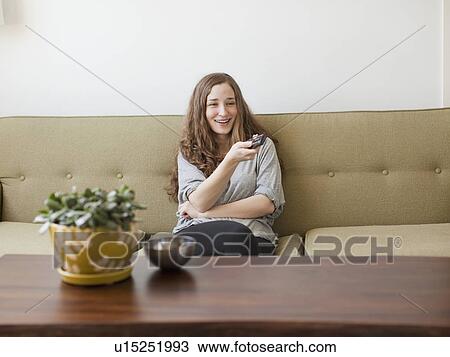 Stock Image - Cheerful young woman sitting on sofa with remote control. Fotosearch