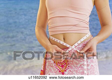 Stock Image - Close up of young woman measuring herself with tape measure, studio shot. Fotosearch