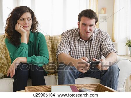 Picture - Couple sitting on sofa, man playing video game. Fotosearch
