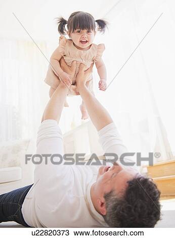 Stock Image - Father lifting baby daughter (12-17 months) while lying on carpet. Fotosearch