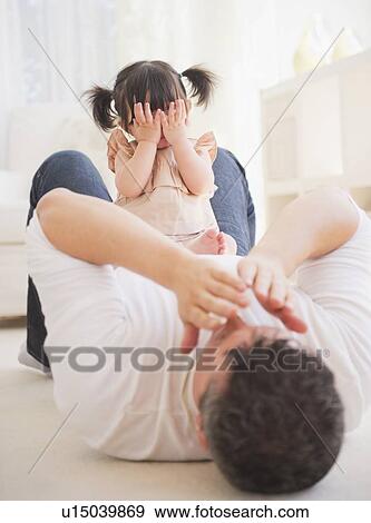 Stock Photo - Father playing with baby daughter (12-17 months) while lying on carpet. Fotosearch