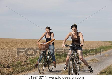 Stock Image - France, Picardie, Albert, Young women on bikes on country road. Fotosearch