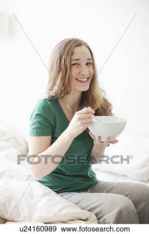 Happy young woman sitting on bed with cornflake bowl View Large Photo Image Stock Photo - Happy young woman sitting on bed with cornflake bowl. Fotosearch