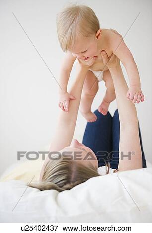 Stock Photo - Mother lying on bed and lifting son (6-11 moths). Fotosearch