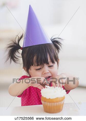 Portrait of baby girl (12-17 months) in party hat eating cupcake View Large Photo Image Stock Photograph - Portrait of baby girl (12-17 months) in party hat eating cupcake. Fotosearch