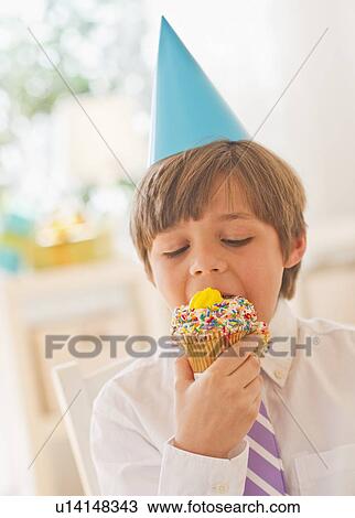 Stock Image - Portrait of boy (10-11 years) in party hat eating cupcake. Fotosearch