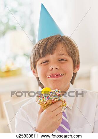 Stock Image - Portrait of boy (10-11 years) in party hat eating cupcake. Fotosearch
