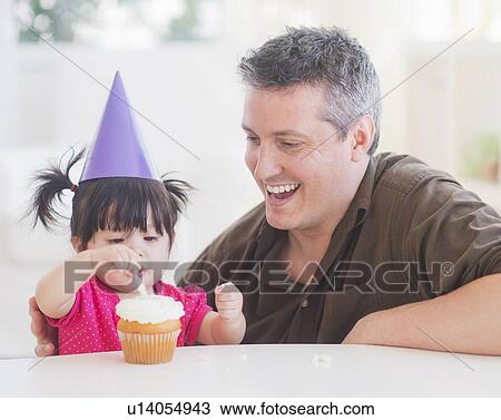 Stock Image - Portrait of father and baby girl (12-17 months) in party hat eating cupcake. Fotosearch