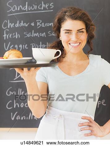 Stock Image - Portrait of smiling waitress with tray against blackboard with menu. Fotosearch