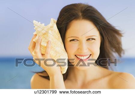 Stock Photo - Portrait of young woman listening to seashell, studio shot. Fotosearch