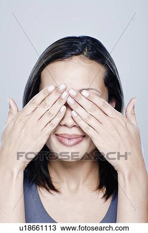 Stock Image - Portrait of young woman with hands covering eyes, studio shot. Fotosearch