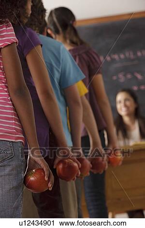 Stock Image - pupils standing in row (10-11, 12-13) holding apple with teacher in background. Fotosearch